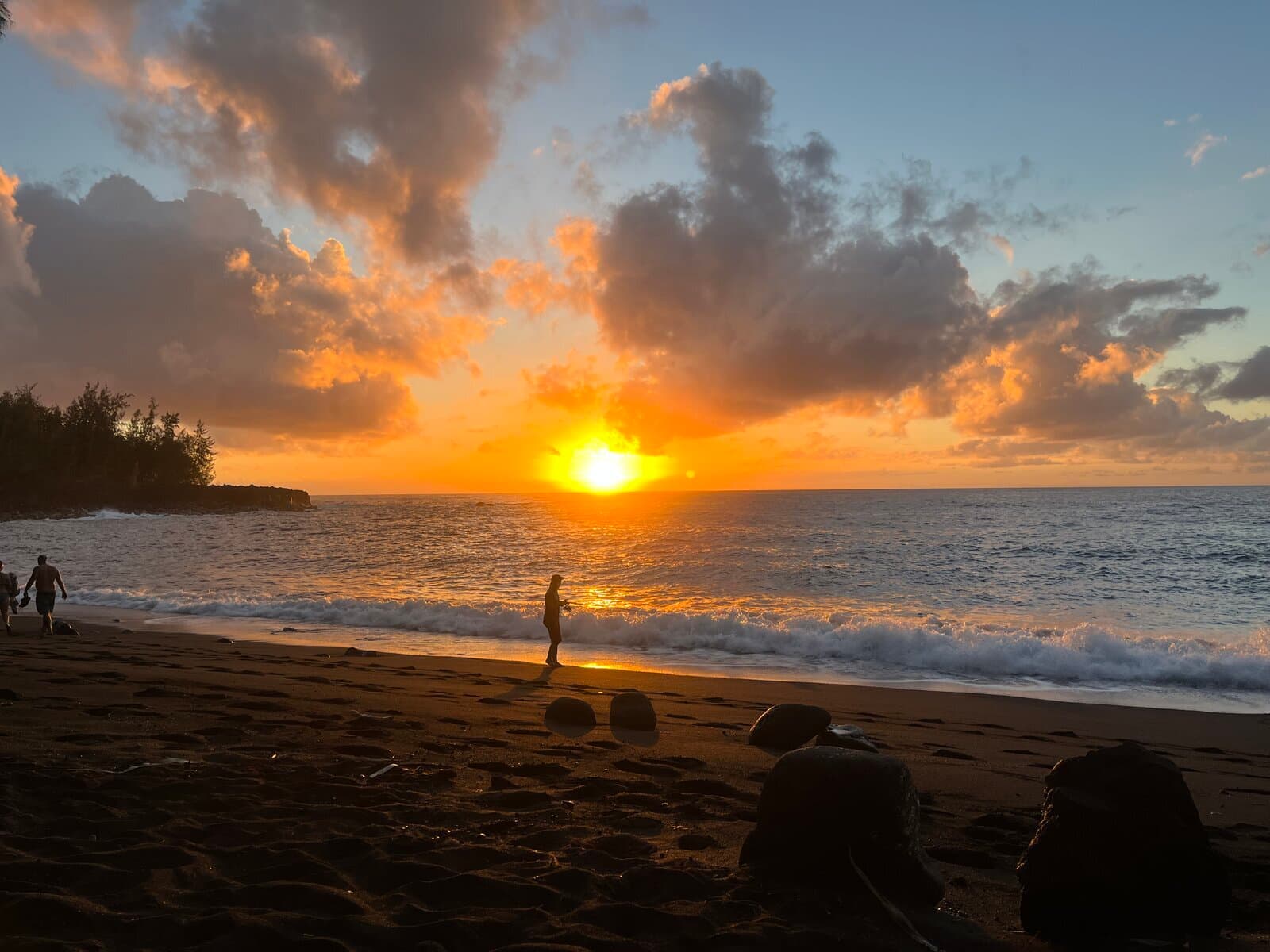 Figures walking a black sand beach as the sun sets over the Big Island coast